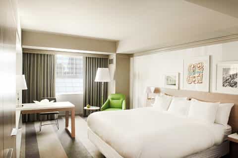 Hotel bedroom with queen bed, desk area, green accent chair, and window with curtains