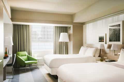 Modern hotel room with two beds, green accent chair, floor-to-ceiling windows, and neutral décor
