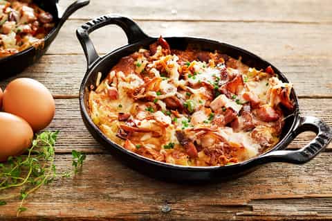 Skillet breakfast dish with eggs, bacon, and cheese on rustic wooden table with fresh herbs