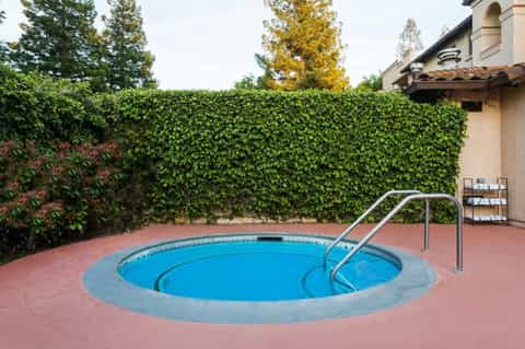 Round hot tub spa surrounded by brick patio, green ivy hedge wall, and metal railings in private resort courtyard