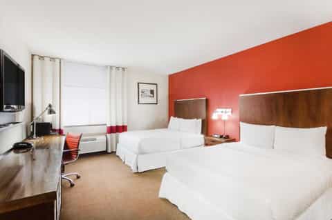 Modern hotel room with two queen beds, coral accent wall, desk area, and flat-screen TV