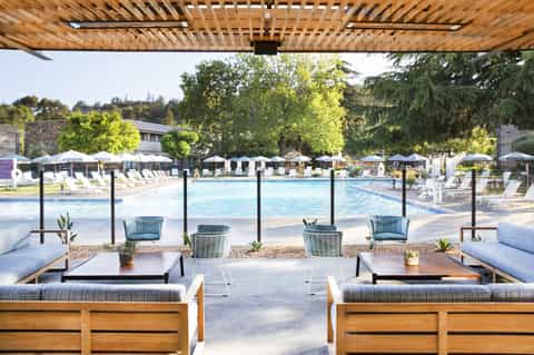 Daytime resort pool scene with wooden pergola, loungers with blue cushions, umbrellas, and trees