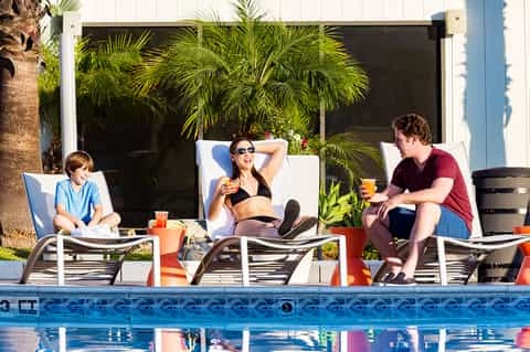 Family relaxing poolside on white loungers with drinks and palm tree backdrop
