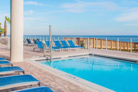Beachfront resort pool with blue loungers, wooden deck, ocean view, and white railings
