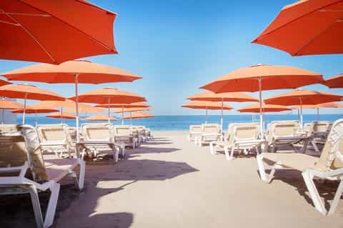 Beachfront lounge with white sun chairs under orange umbrellas facing the ocean and blue sky