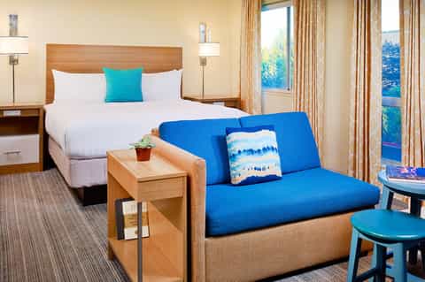Hotel guest room with queen bed, blue sofa, and balcony overlooking trees
