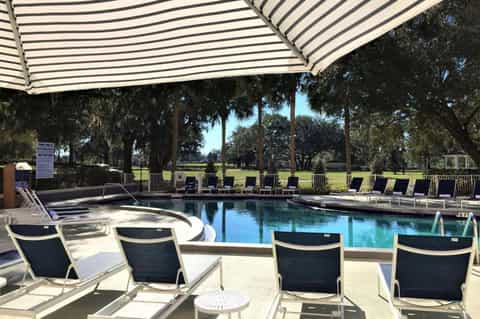 Resort pool with striped umbrellas, lounge chairs, and views of manicured grounds with large oak trees