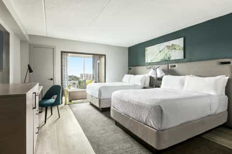 Hotel guest room with two queen beds, teal accent wall, desk, and balcony with city views