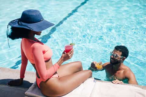 Woman in pink swimwear and hat holding cocktail by pool edge while man swims nearby