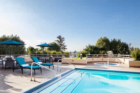 Resort pool with lane markers, lounge chairs with blue cushions, umbrellas, and scenic garden views