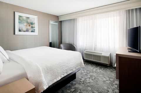 Modern hotel bedroom with white bedding, desk area, and floor-to-ceiling windows