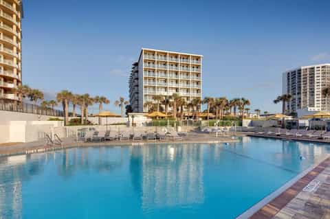 Large outdoor resort pool with lounge chairs, palm trees, and beachfront buildings