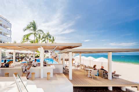 Beachfront resort bar and lounge overlooking ocean with palm trees and white umbrellas
