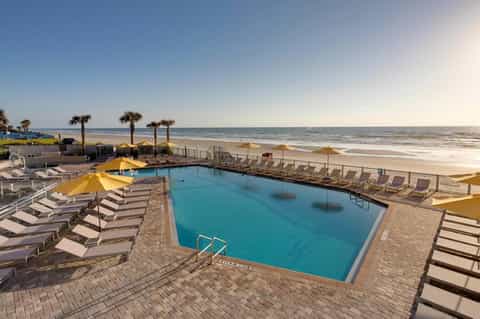 Beachfront resort pool with yellow umbrellas, lounge chairs, sandy beach, and ocean horizon