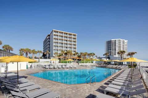 Beachfront resort pool with yellow umbrellas, high-rise buildings, and palm trees under clear blue sky