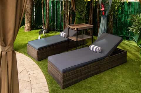 Luxury resort spa area with rattan loungers, gray cushions, tropical plants, green gates, and curtained cabanas on grass