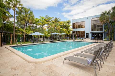 Rectangular pool surrounded by palm trees, blue umbrellas, modern white building