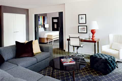 Modern hotel suite living room with gray sofa, desk, and view to bedroom