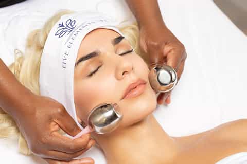 Spa facial treatment with therapist applying metal cooling spheres to client's face wearing headband