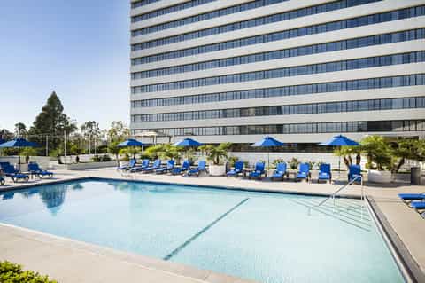 Large rectangular pool with blue umbrellas and loungers against modern high-rise hotel building