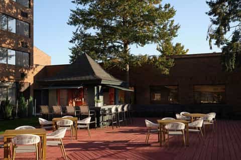Outdoor patio with woven chairs, wooden tables, brick building, and mature shade trees overhead