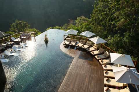 Infinity pool overlooking forested hillside with wooden deck, lounge chairs, and white umbrellas