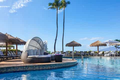 Luxurious resort pool with cabana, palm trees, tiki umbrellas, and ocean backdrop