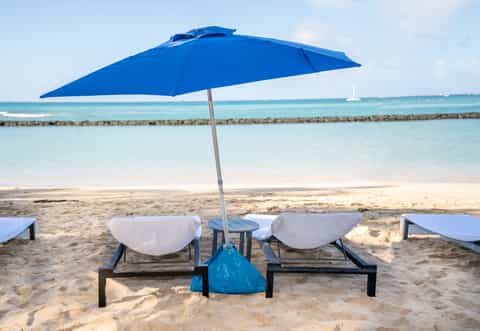 Two beach lounge chairs with white cushions under blue umbrella facing calm turquoise ocean