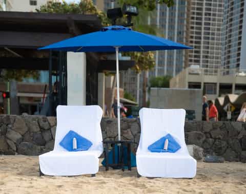 Two white pool loungers under blue umbrella on urban beach with city skyline