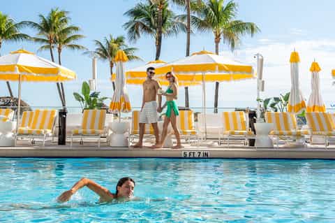 Resort pool with yellow and white umbrellas, swimmers, and palm trees against ocean view