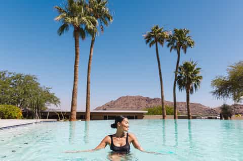 Woman in pool with palm trees and desert mountain backdrop under clear blue sky