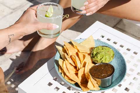Hands holding margarita with chips and guacamole on blue plate outdoors
