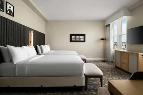 Contemporary hotel room with two beds, dark headboard, and city window views