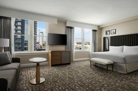 Hotel bedroom with king bed, black headboard, city views through floor-to-ceiling windows, and seating area