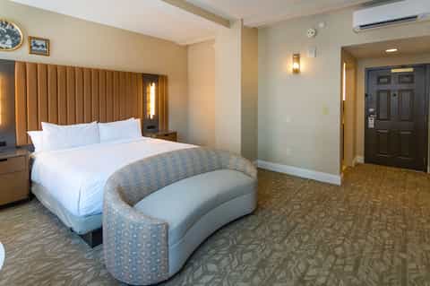 Modern hotel room with queen bed, padded headboard, curved seating, and neutral décor tones