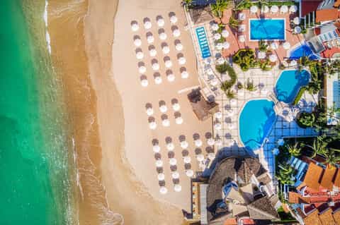 Aerial view of beachfront resort with arranged umbrellas, blue pools, sandy beach, and turquoise ocean