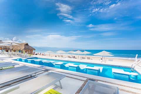 Bright blue resort pool with geometric white sun loungers overlooking pristine beach and turquoise ocean