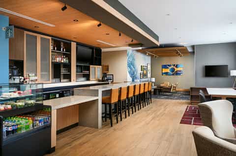 Modern hotel bar and lounge with wooden ceiling, orange bar stools, display shelving, beverage coolers, and seating area