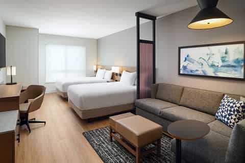 Hotel guest room with two queen beds, gray sofa, work desk, and warm contemporary decor