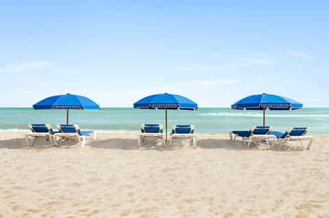 Beach scene with three blue umbrellas, white lounge chairs, turquoise ocean, and sandy shore