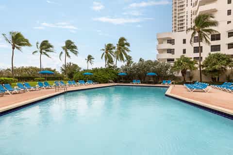 Large rectangular resort pool with blue lounge chairs and umbrellas, flanked by palm trees and beige buildings