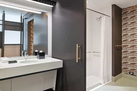 Modern hotel bathroom with white sink, dark wall panels, glass shower enclosure, and decorative tile accent