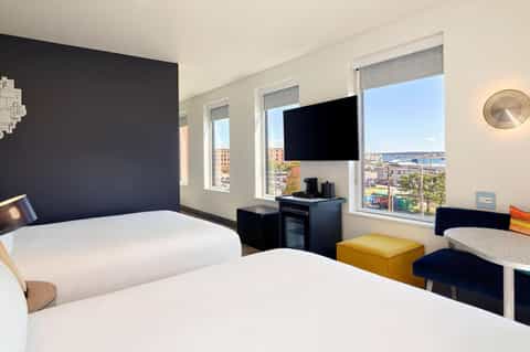 Modern hotel room with two beds, dark accent wall, flat-screen TV, and ocean city view windows