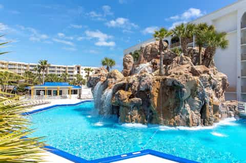 Resort pool with rocky waterfall feature, blue water, palm trees, and multi-story accommodations