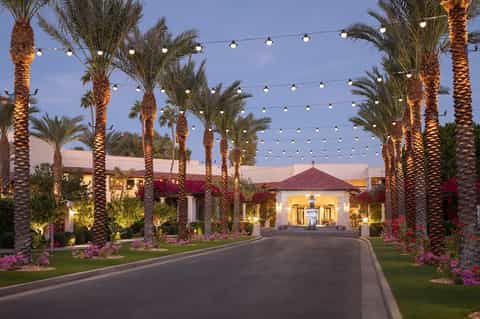 Palm-lined driveway lit by string lights and uplighting, leading to illuminated Spanish-style villa entrance at dusk