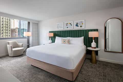 Modern hotel bedroom with green velvet headboard, white bedding, wood furniture, and city views