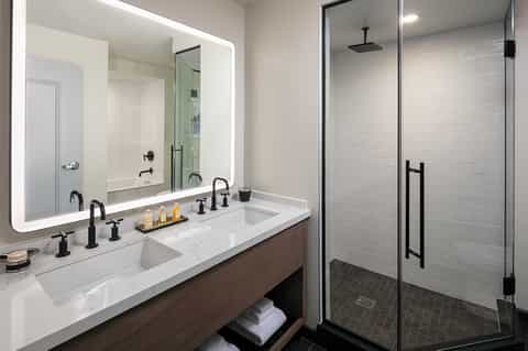 Contemporary bathroom with double vanity, illuminated mirror, white marble counters, and walk-in shower