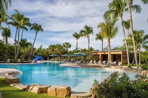 Large resort pool surrounded by palm trees, lounge chairs, cabanas, and manicured lawns under blue sky