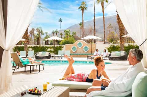 Couple relaxing by turquoise pool under white cabana with mountain views and palm trees