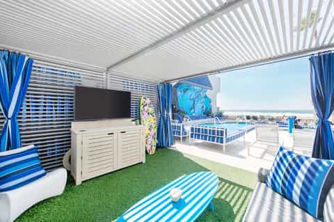 Covered beach cabana with blue striped furnishings, TV, and ocean view beyond railing